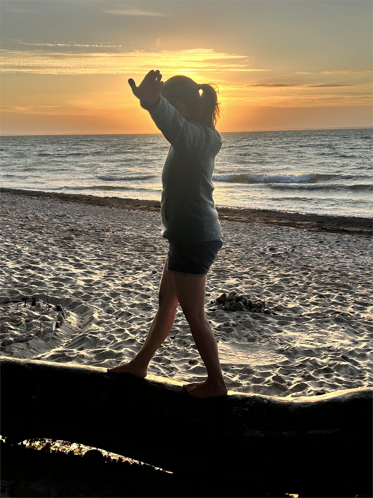 Ein Kind balanciert barfuß über einen Baumstamm am Strand, dahinter geht die Sonne über dem Meer unter.
