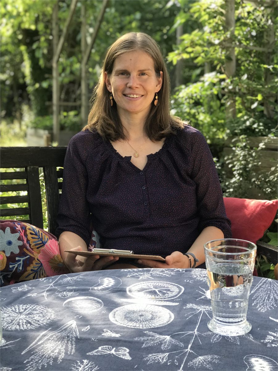 Nadine Kreuzer sitzt an einem Gartentisch, vor sich ein Klemmbrett und ein Glas Wasser.