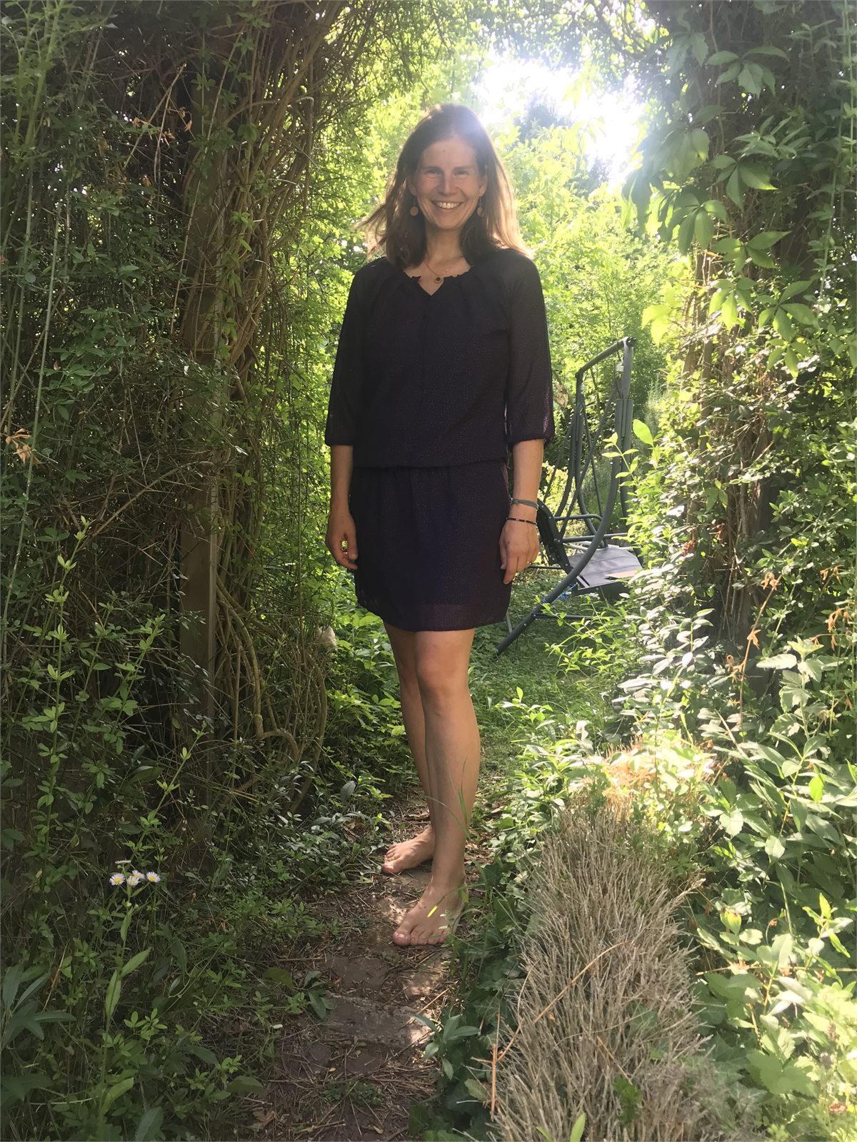Nadine Kreuzer auf einem sonnendurchfluteten Gartenweg zwischen hohen grünen Hecken.
